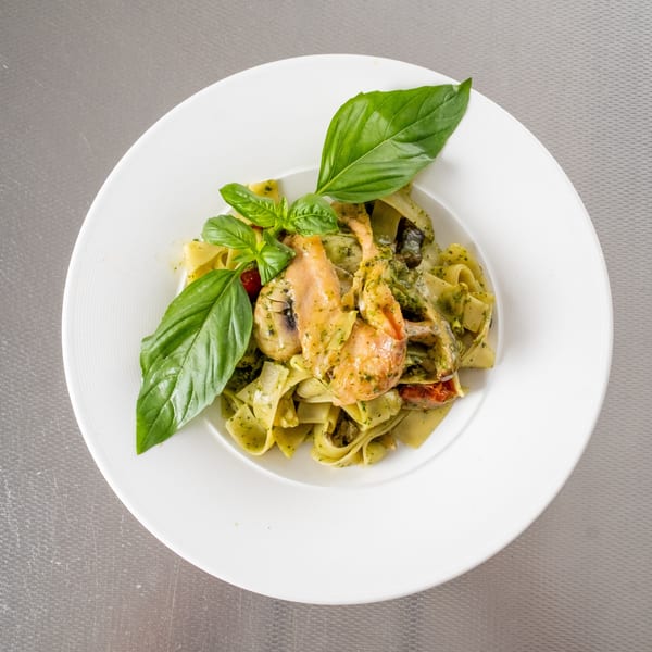 White plate of pesto pasta with salmon, mushrooms, tomatoes, and fresh basil.