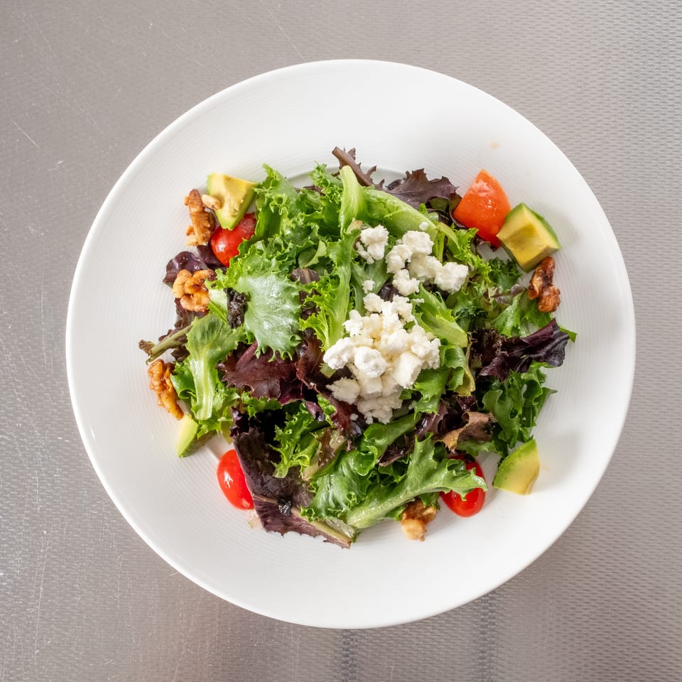 Fresh mixed green salad with tomatoes, avocado, walnuts, and feta cheese in a white bowl.