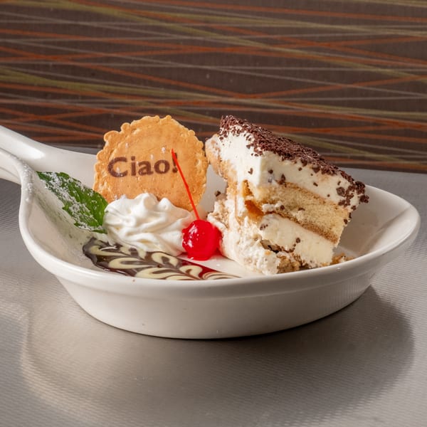 Slice of tiramisu with whipped cream, cherry, mint, and a "Ciao!" cookie on a white dish.
