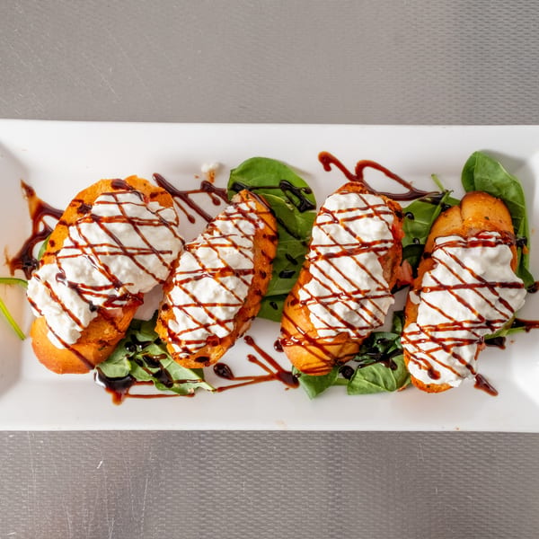 Four ricotta crostini drizzled with balsamic glaze, served on spinach leaves on a white plate.