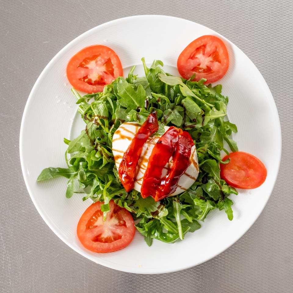 Arugula salad with mozzarella, roasted red peppers, tomatoes, and balsamic glaze on a white plate.