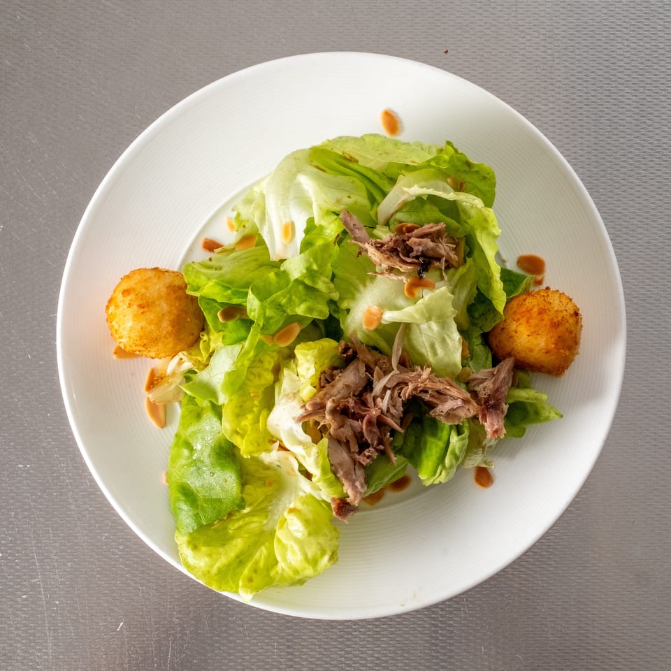 Fresh green lettuce salad with shredded meat, two fried croquettes, and dressing on a white plate.