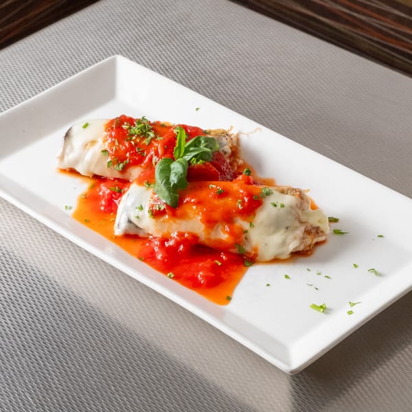 Two pasta rolls covered in red sauce, melted cheese, and fresh basil on a white rectangular plate.