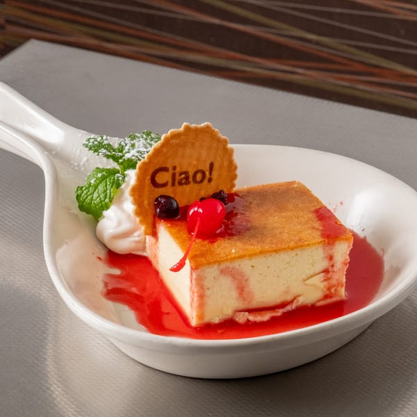 Square dessert with red sauce, whipped cream, mint, cherry, and a "Ciao!" wafer on a white plate.